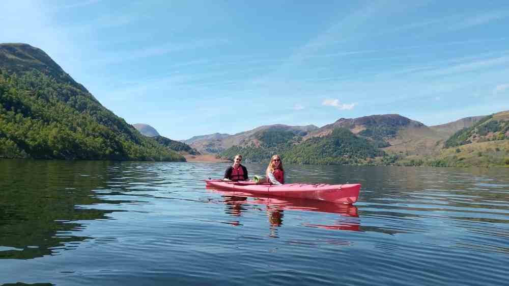 A couple kayaking together on a calm lake in the Lake District, surrounded by rolling hills and beautiful blue skies.