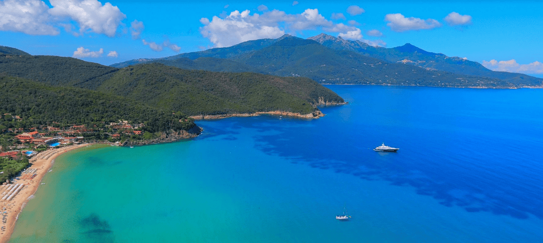 Aerial view of a beach, hills, and boats on clear blue water under a sunny sky.