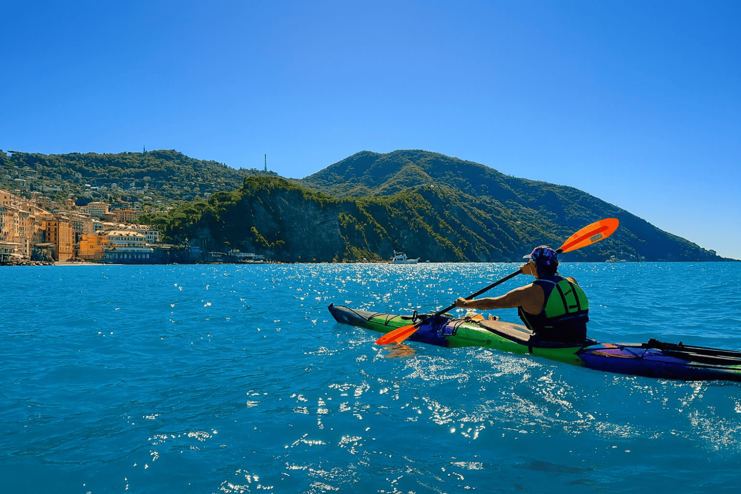 Person kayaking on blue sea near coastal town with hills in the background.