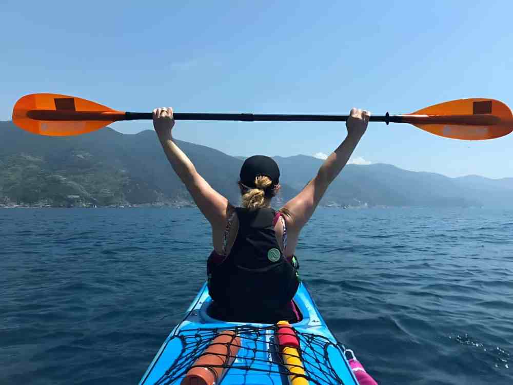 Kayaking along the Italian coast Woman kayaking, holding a paddle overhead on a calm sea with mountains in the background.
