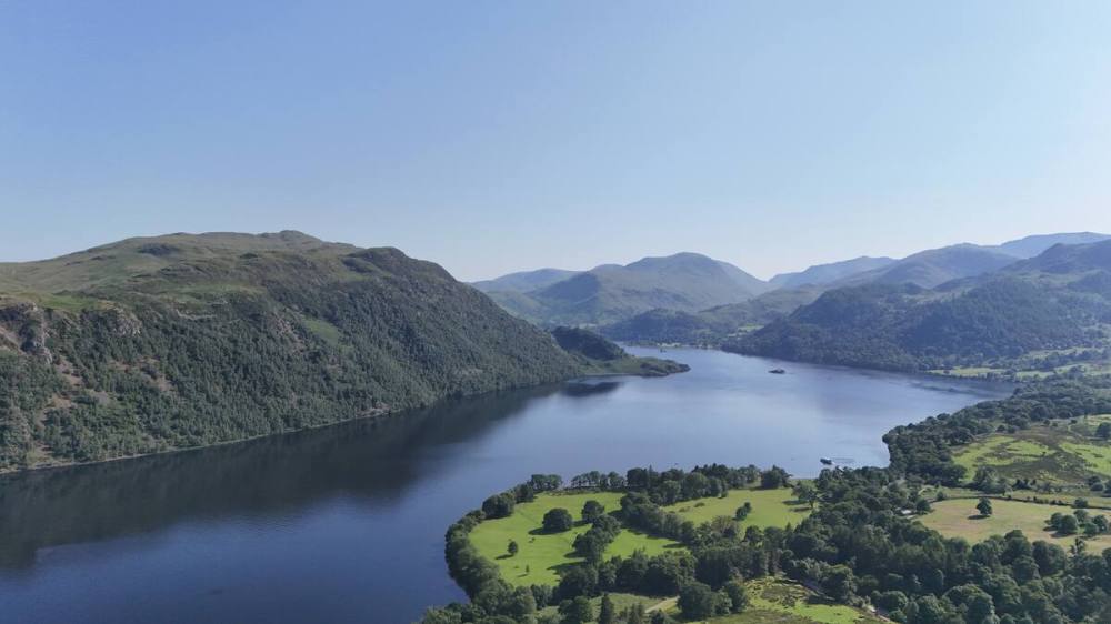 Aerial view of a serene lake surrounded by green hills and mountains under a clear blue sky.