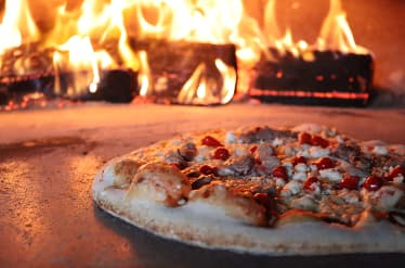 Wood-fired pizza baking in a brick oven with visible flames.