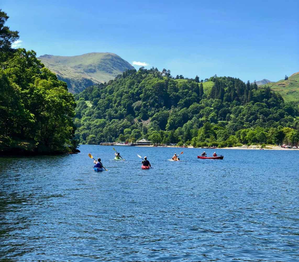 Kayaking along stunning tree lined shores. People kayaking on a lake surrounded by lush green hills under a clear blue sky.