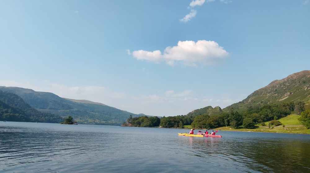 Ullswater Place Names Two people kayaking on a lake surrounded by mountains under a blue sky with a few clouds.
