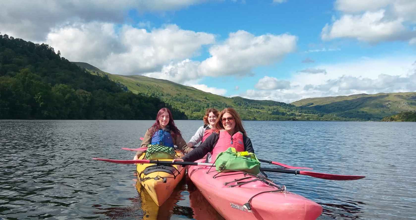 Three people kayaking on a lake with hills and clouds in the background.