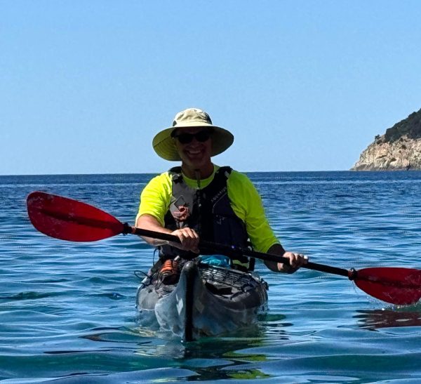 Person kayaking on clear blue sea near rocky coastline, wearing a hat and neon shirt.