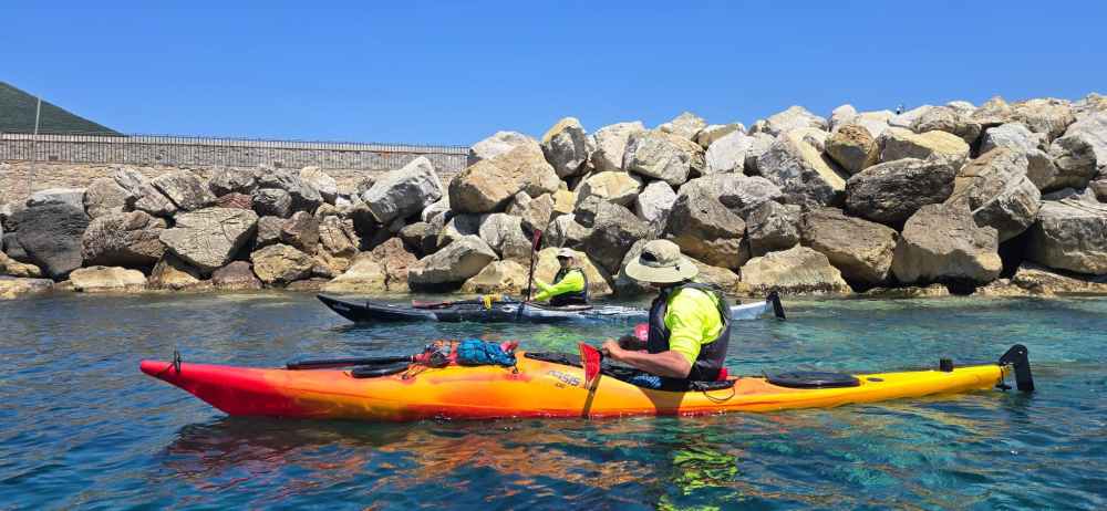 Two kayakers in bright clothing paddle near rocky shoreline.