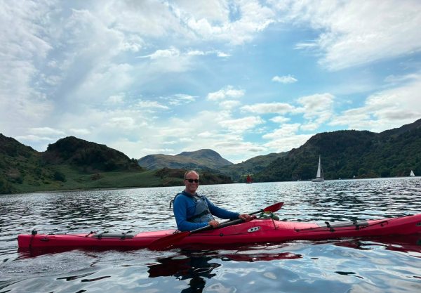 Person kayaking on a lake with mountains and sailboats in the background.